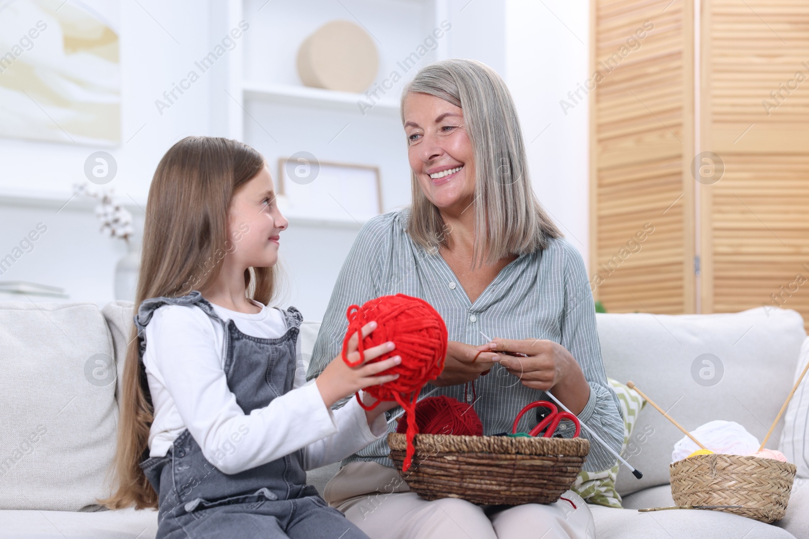 Photo of Smiling grandmother teaching her granddaughter to knit on sofa at home