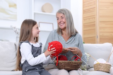 Smiling grandmother teaching her granddaughter to knit on sofa at home