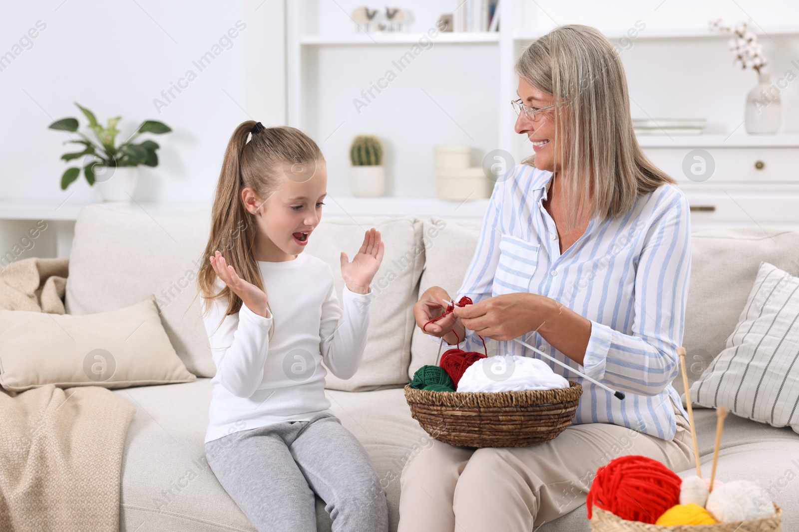 Photo of Smiling grandmother teaching her granddaughter to knit on sofa at home