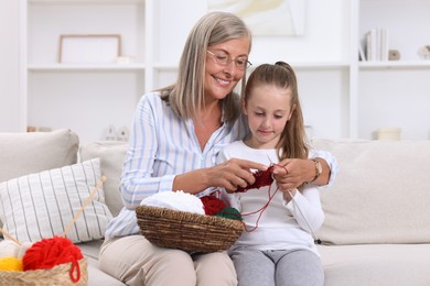 Photo of Smiling grandmother teaching her granddaughter to knit on sofa at home