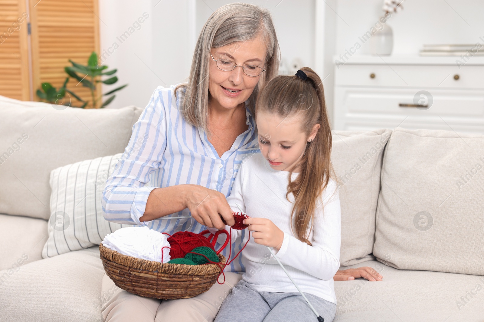 Photo of Smiling grandmother teaching her granddaughter to knit on sofa at home