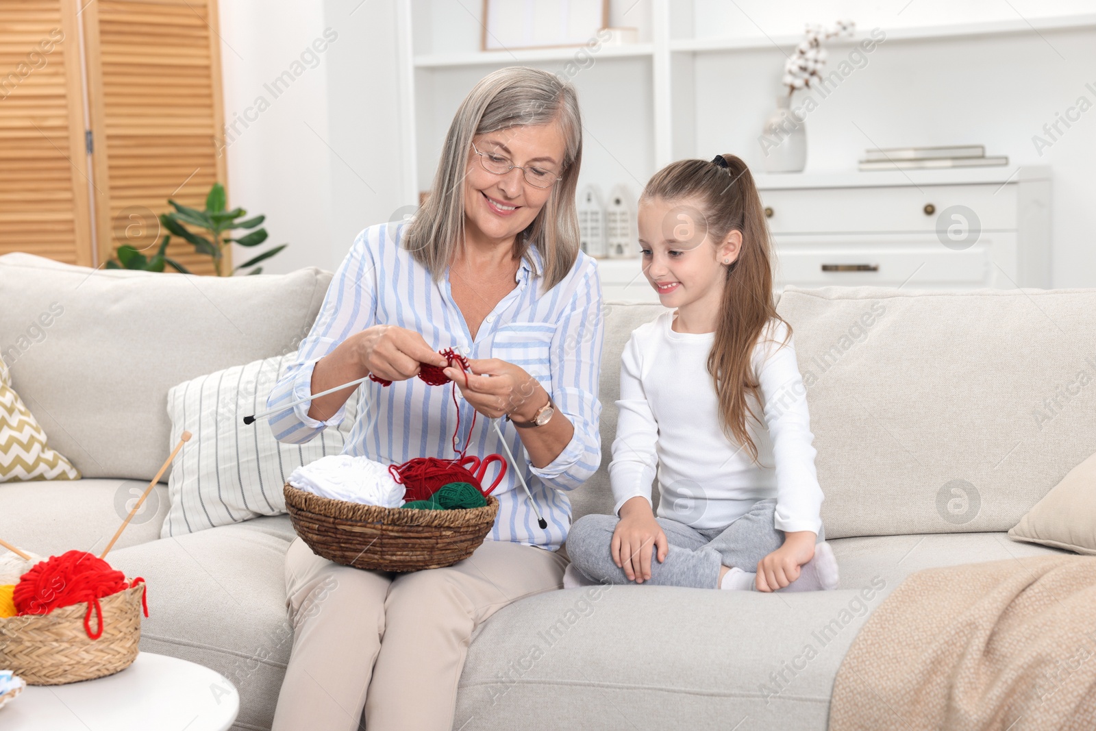 Photo of Smiling grandmother teaching her granddaughter to knit on sofa at home