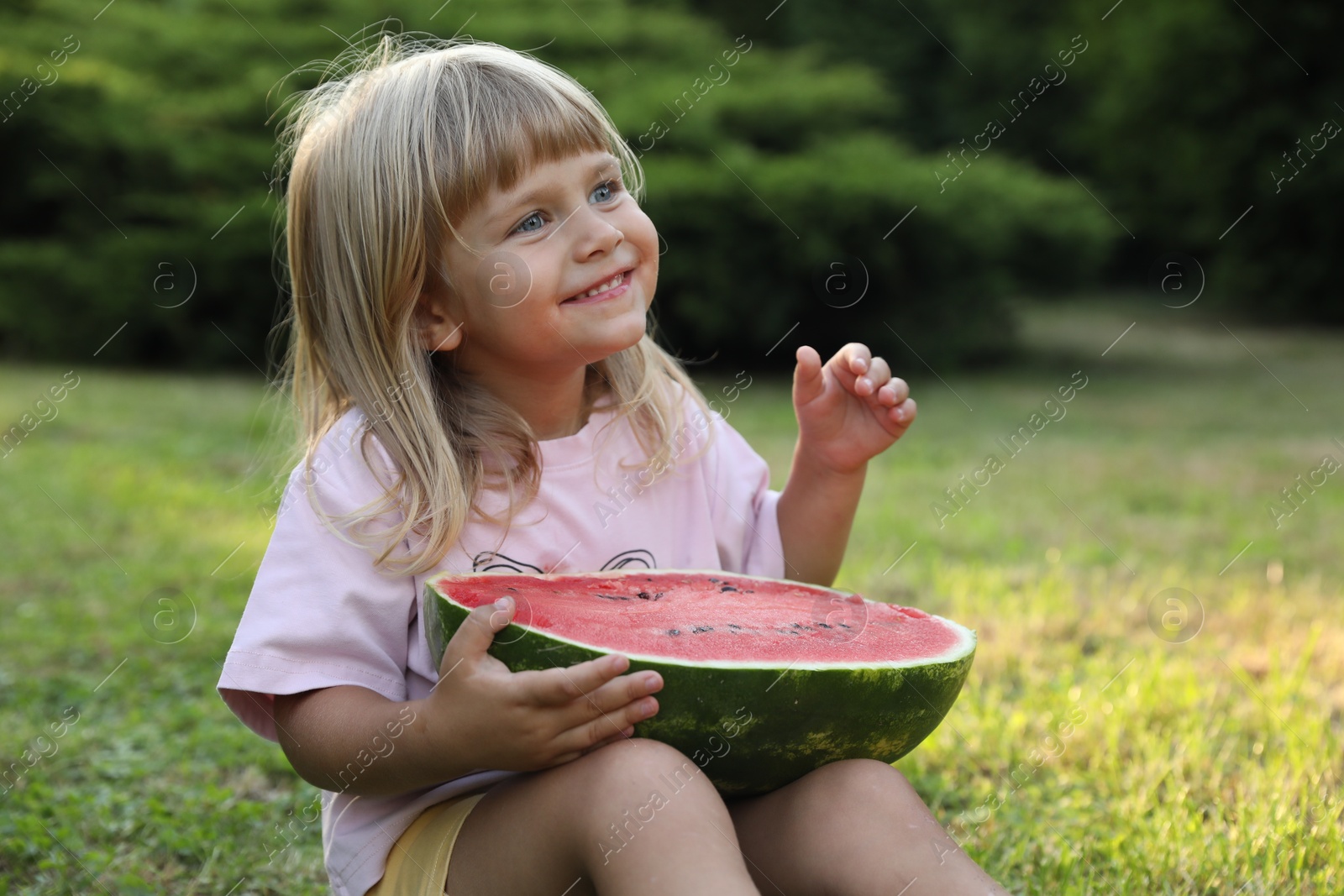 Photo of Cute little girl with half of juicy watermelon on green grass outdoors