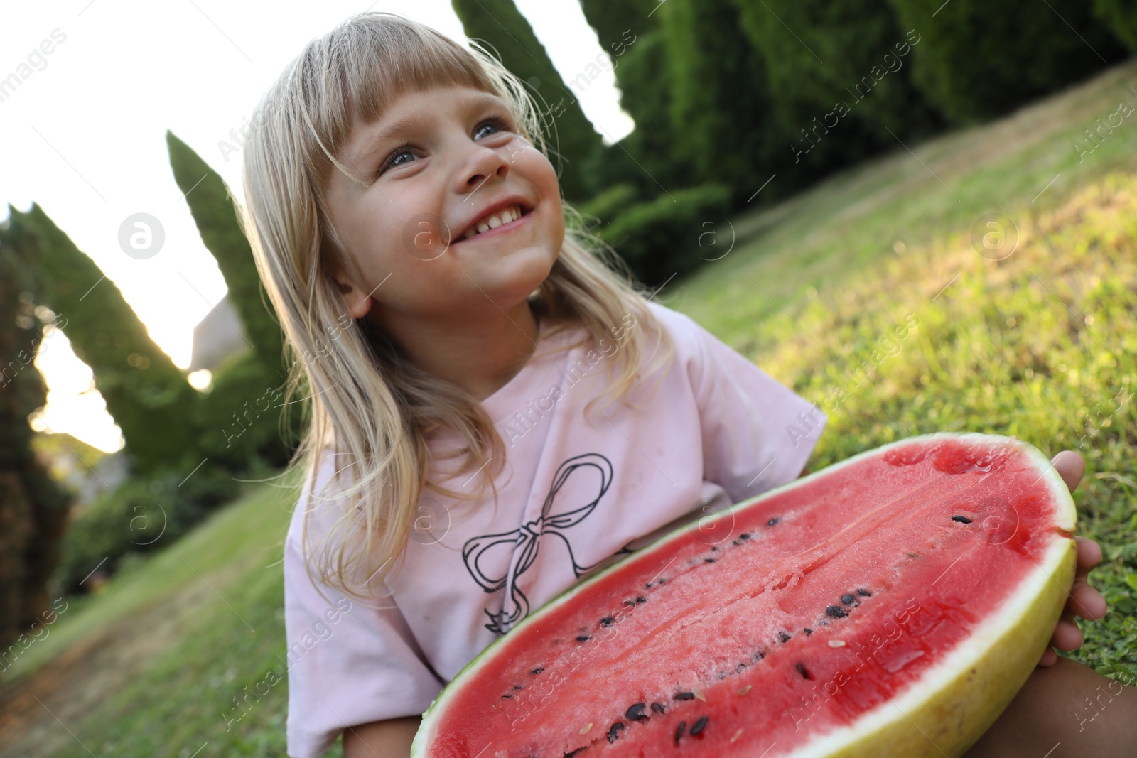 Photo of Cute little girl with half of juicy watermelon on green grass outdoors