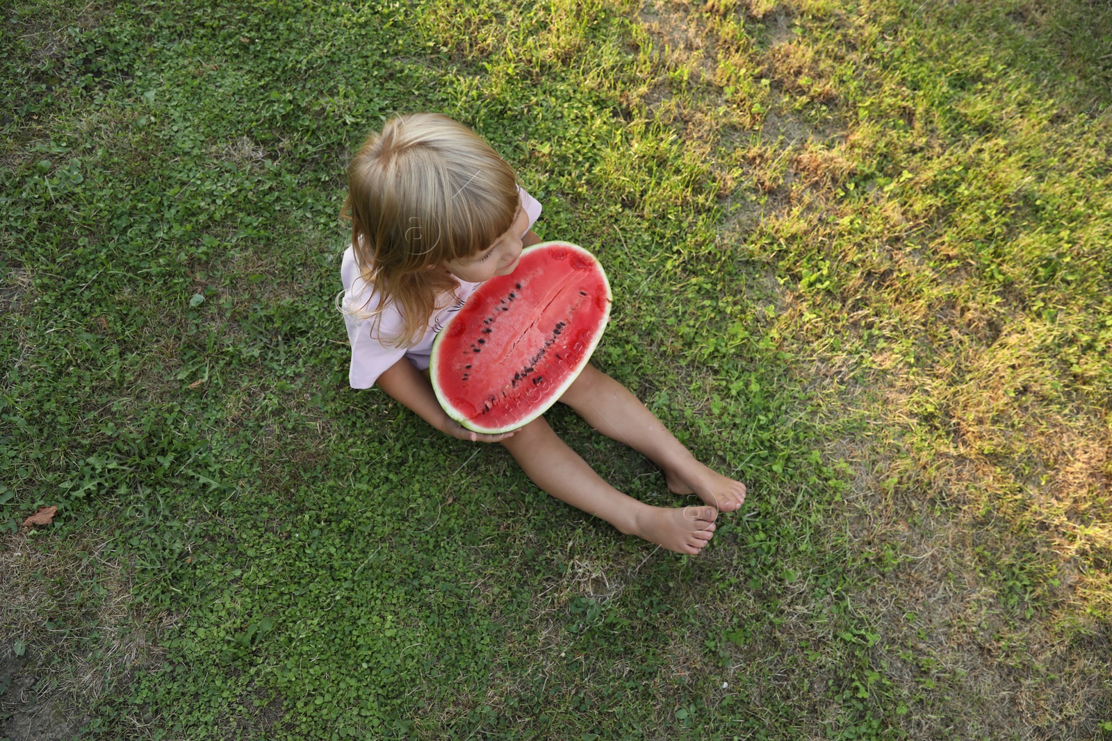 Photo of Cute little girl with half of juicy watermelon on green grass outdoors, above view