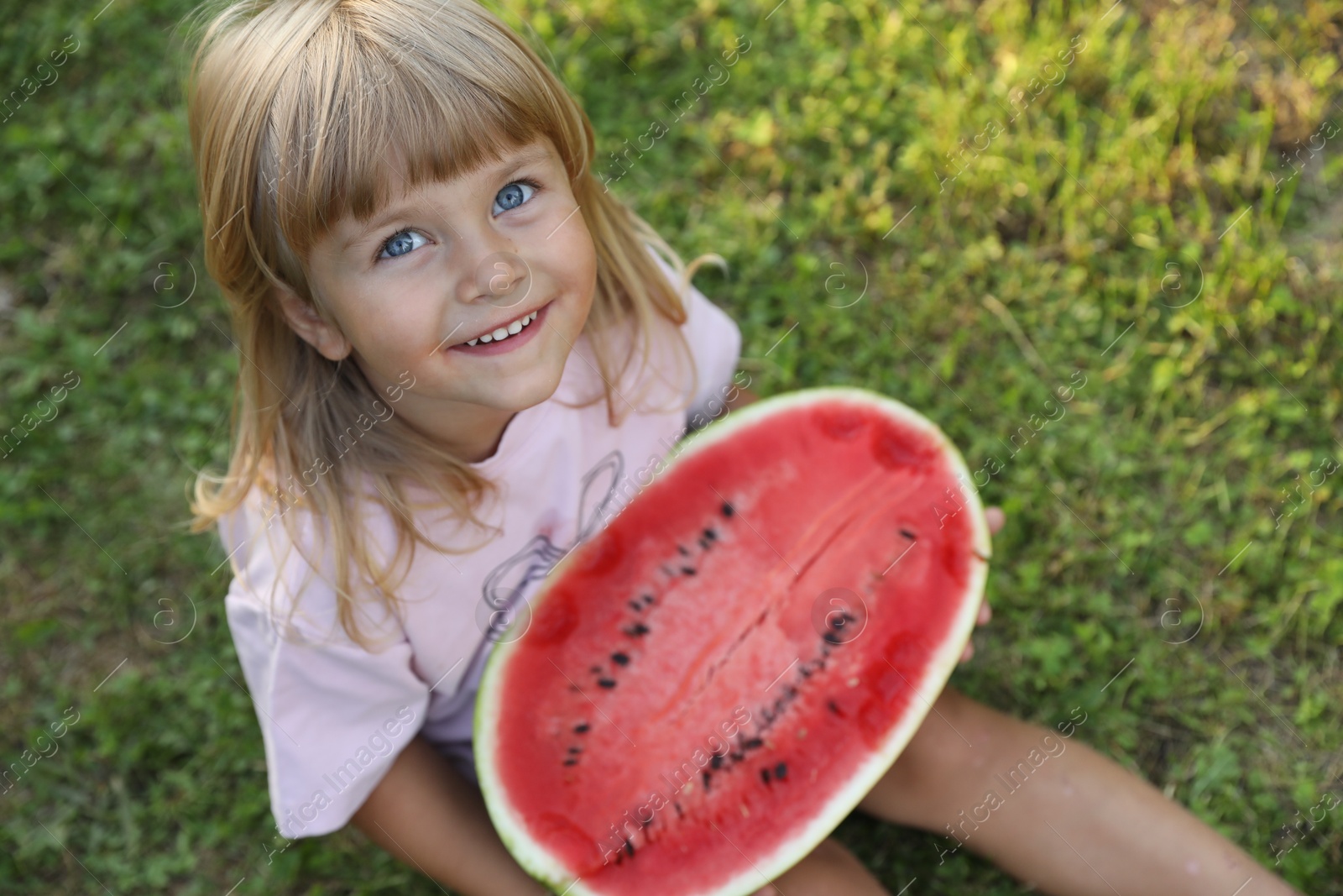 Photo of Cute little girl with half of juicy watermelon on green grass outdoors, above view