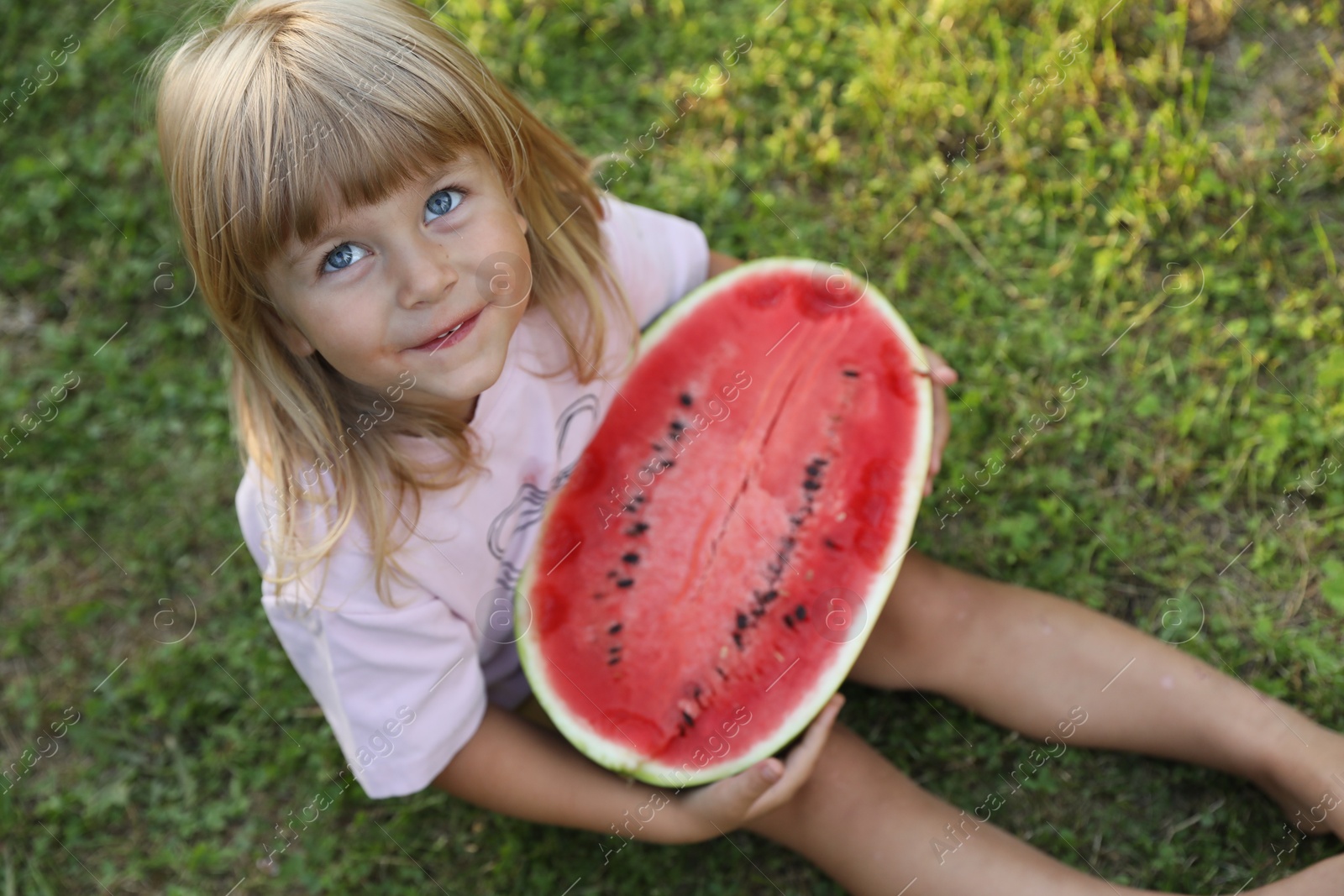 Photo of Cute little girl with half of juicy watermelon on green grass outdoors, above view