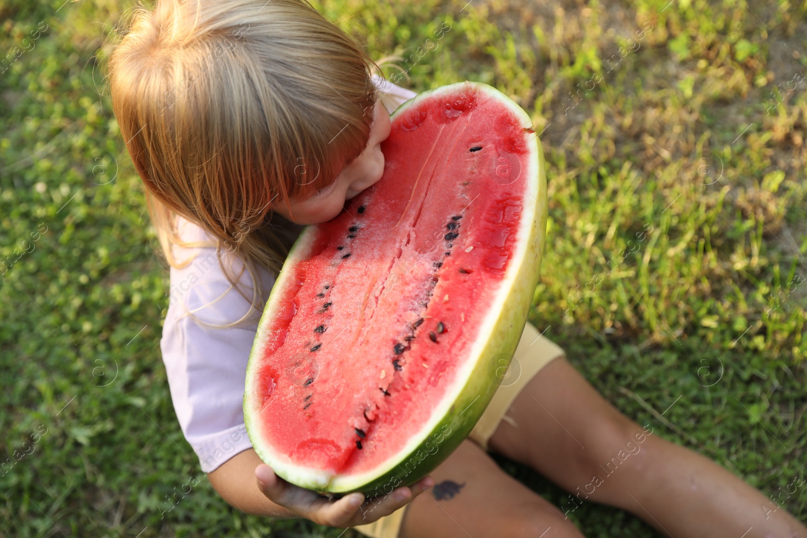 Photo of Cute little girl with half of juicy watermelon on green grass outdoors, above view