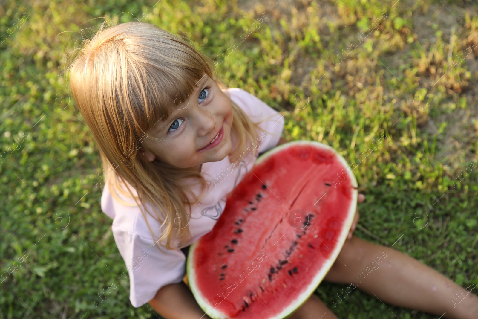 Photo of Cute little girl with half of juicy watermelon on green grass outdoors, above view