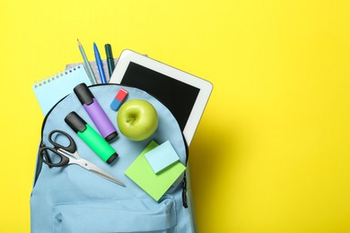 Photo of Light blue school backpack with stationery, apple and tablet on yellow background, top view. Space for text