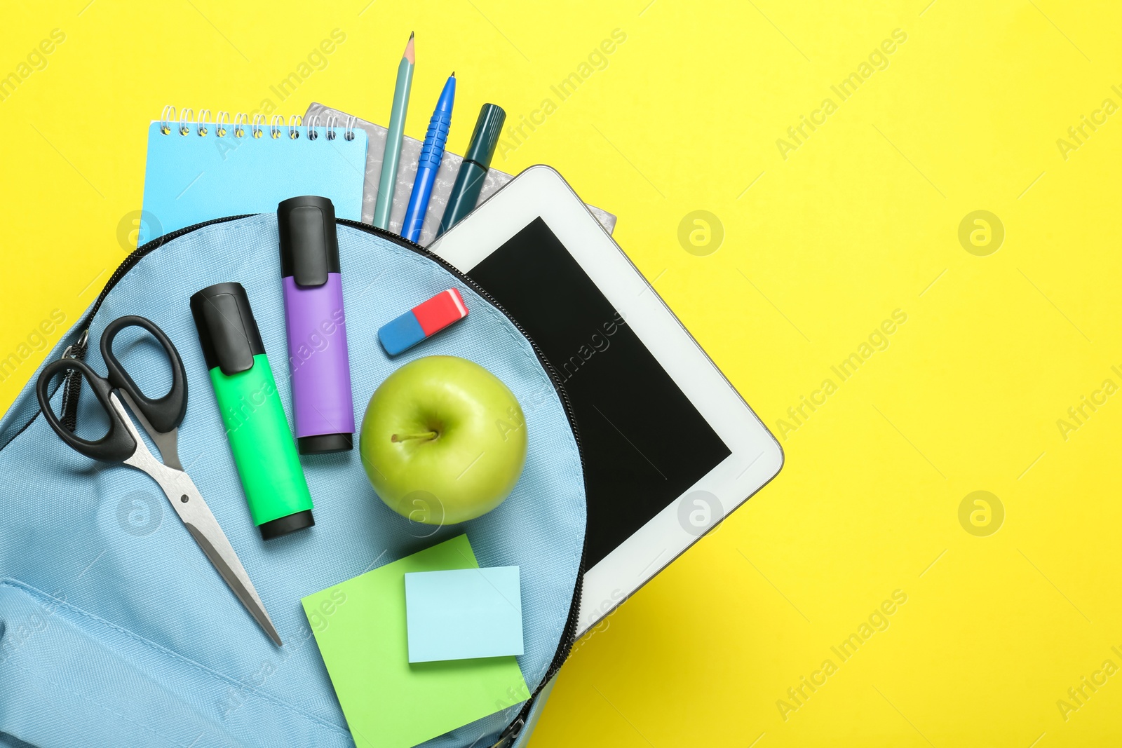 Photo of Light blue school backpack with stationery, apple and tablet on yellow background, top view. Space for text