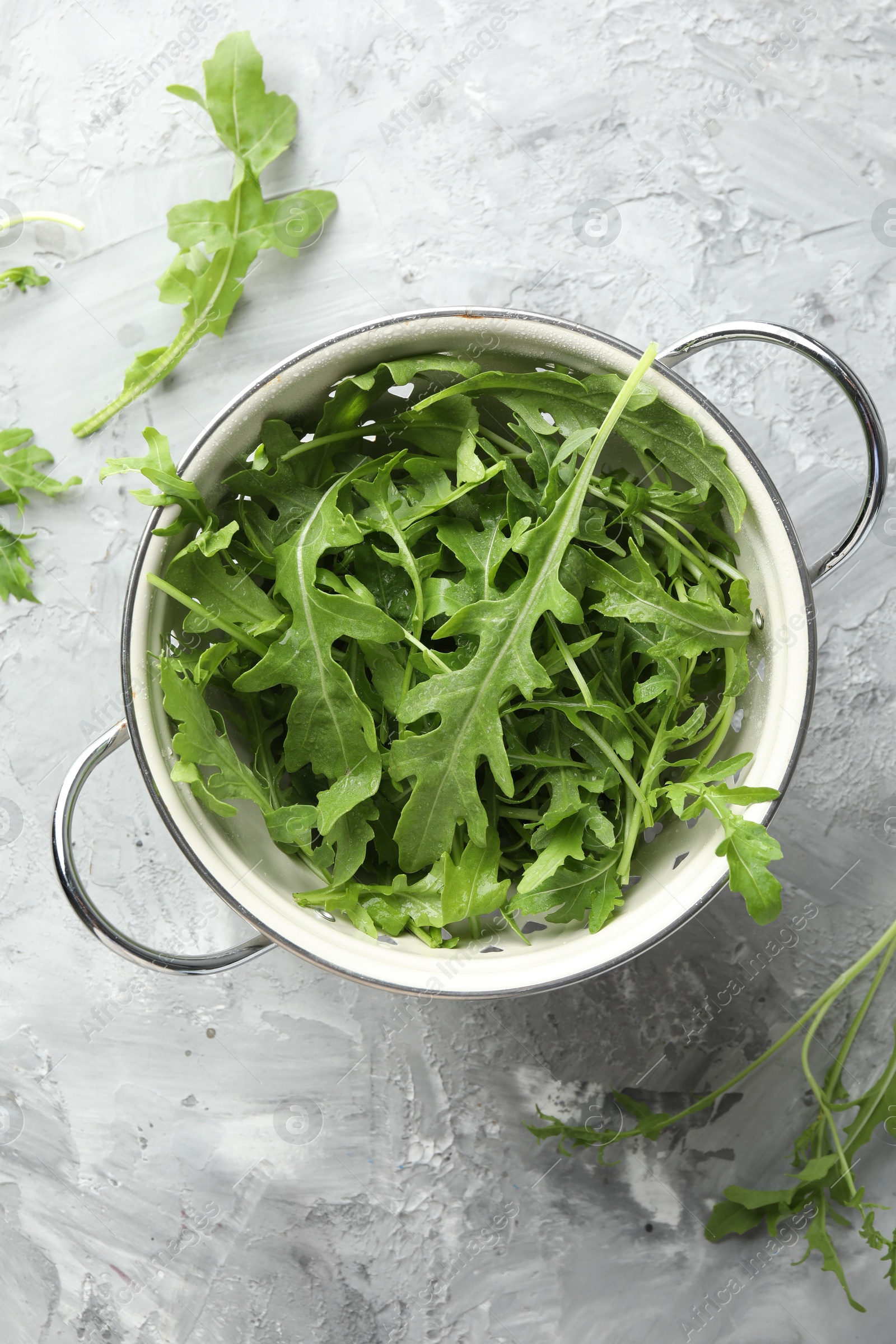 Photo of Fresh green arugula leaves in colander on grey textured table, top view