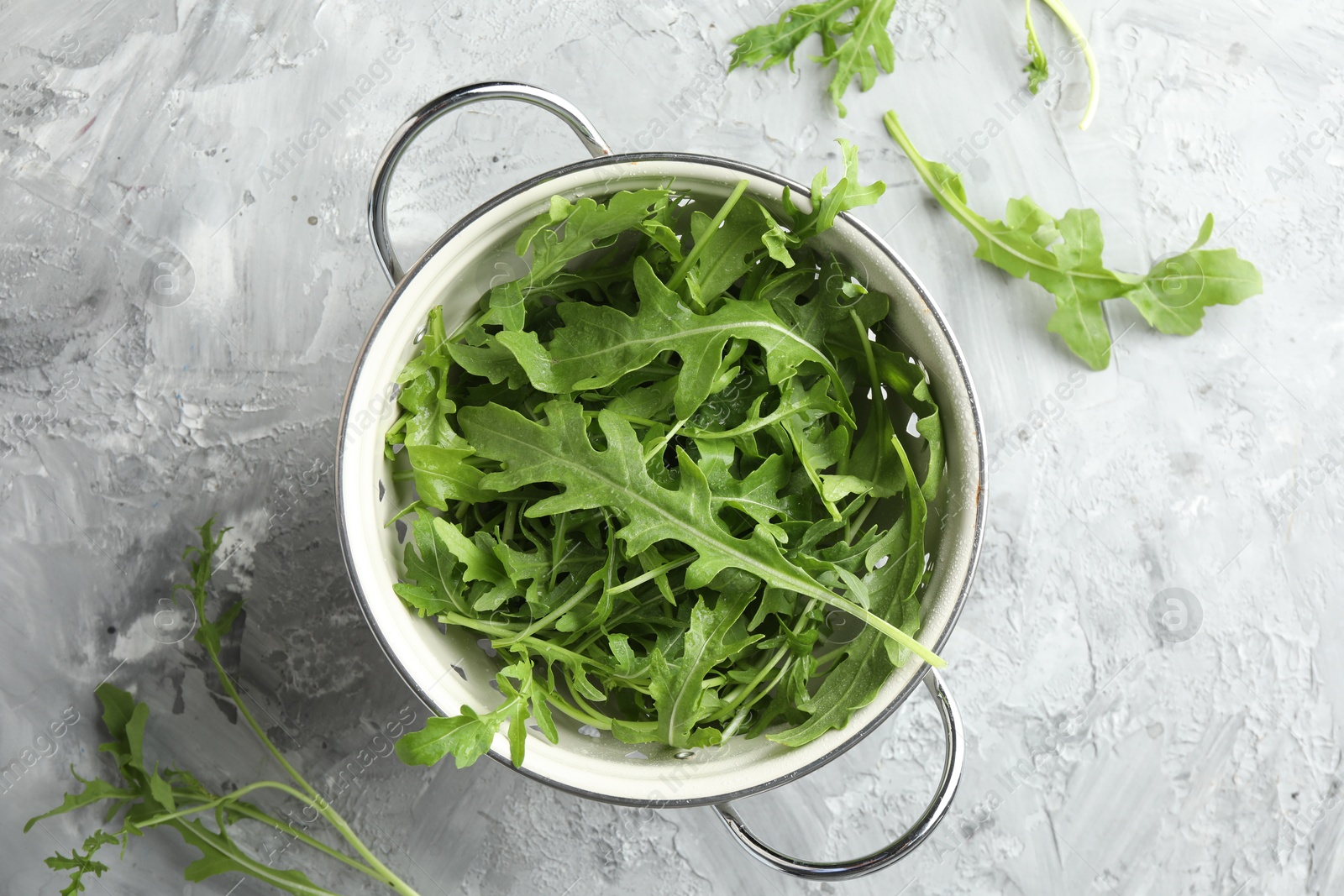 Photo of Fresh green arugula leaves in colander on grey textured table, top view