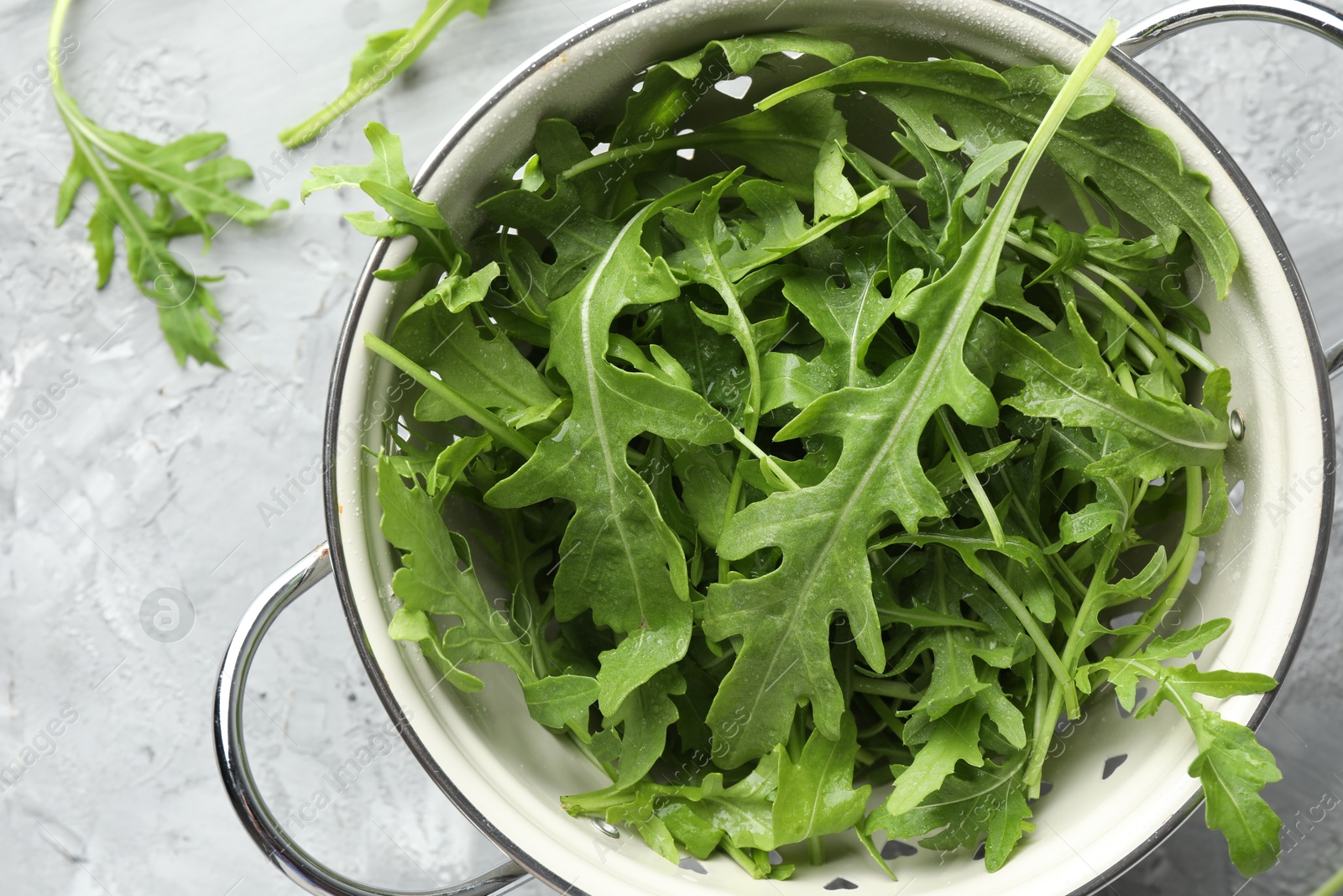 Photo of Fresh green arugula leaves in colander on grey textured table, top view