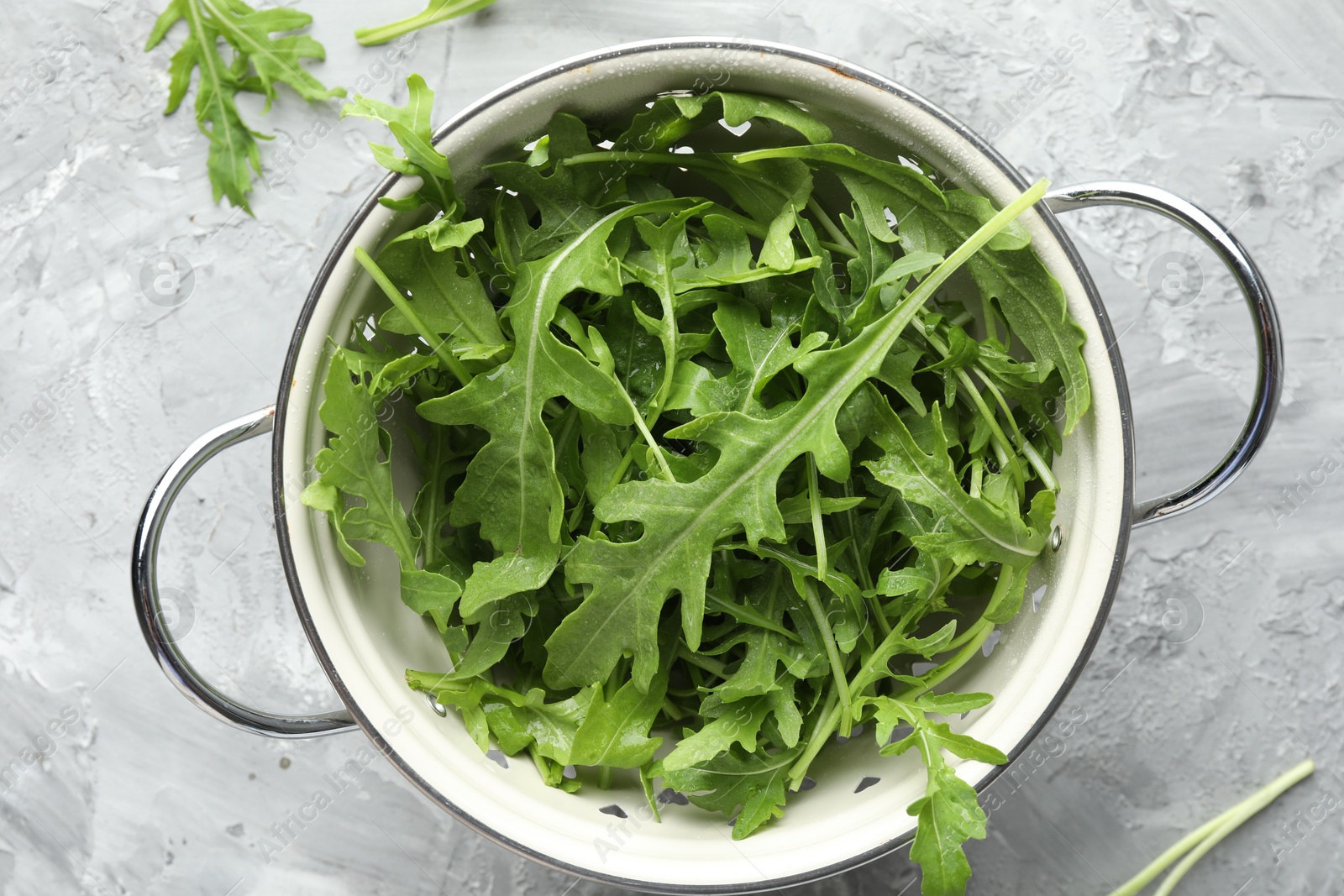 Photo of Fresh green arugula leaves in colander on grey textured table, top view