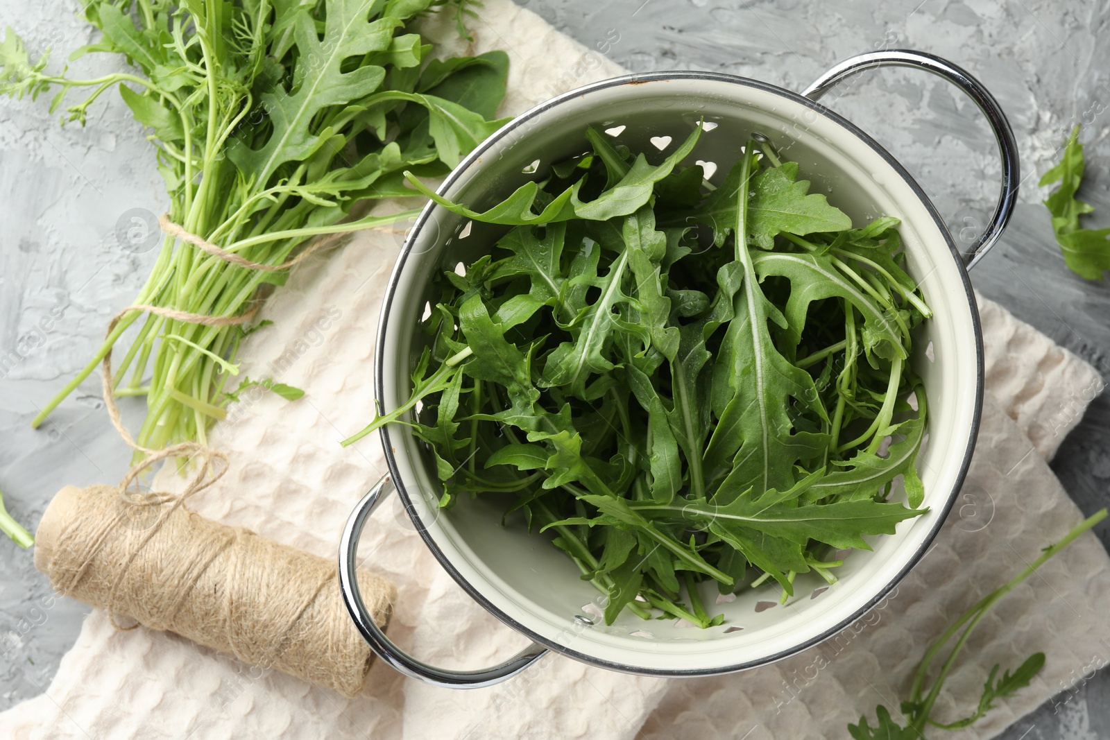 Photo of Fresh green arugula leaves in colander and spool of thread on grey table, flat lay