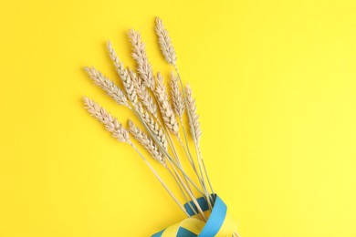 Photo of Ears of wheat with ribbon in colors of Ukrainian national flag on yellow background, top view