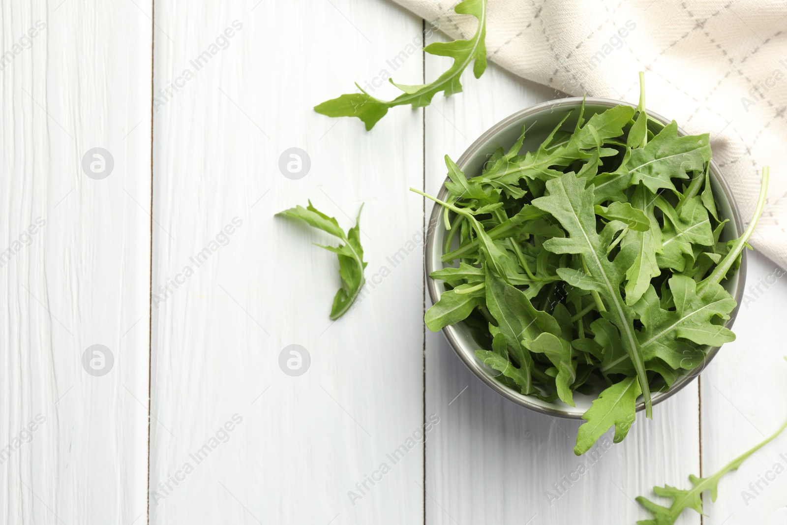 Photo of Fresh green arugula leaves in bowl on white wooden table, top view. Space for text