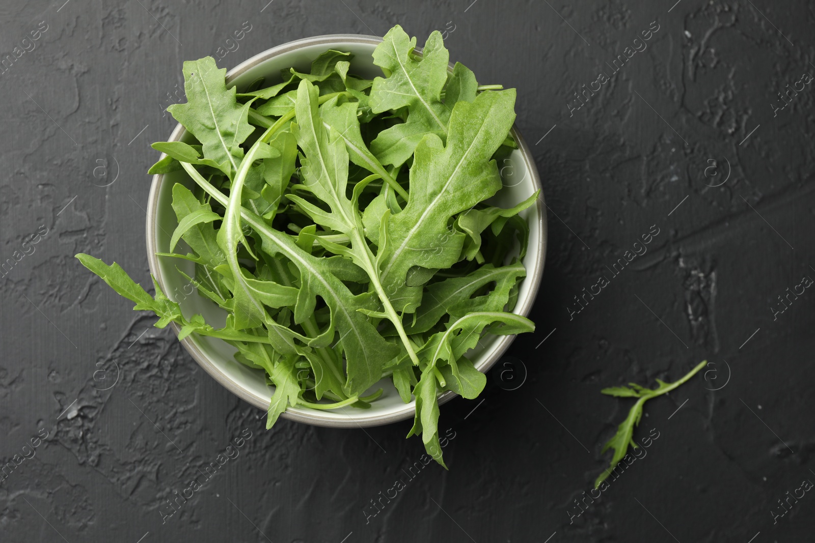 Photo of Fresh green arugula leaves in bowl on grey textured table, top view