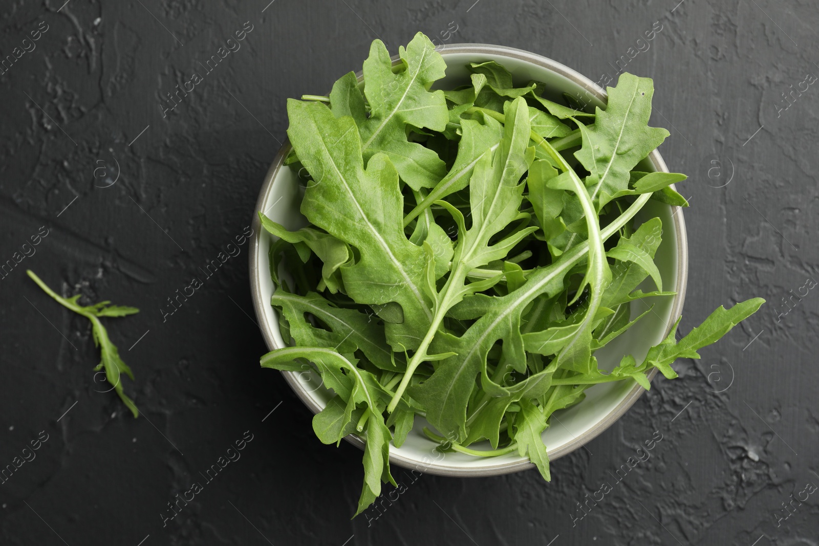 Photo of Fresh green arugula leaves in bowl on grey textured table, top view