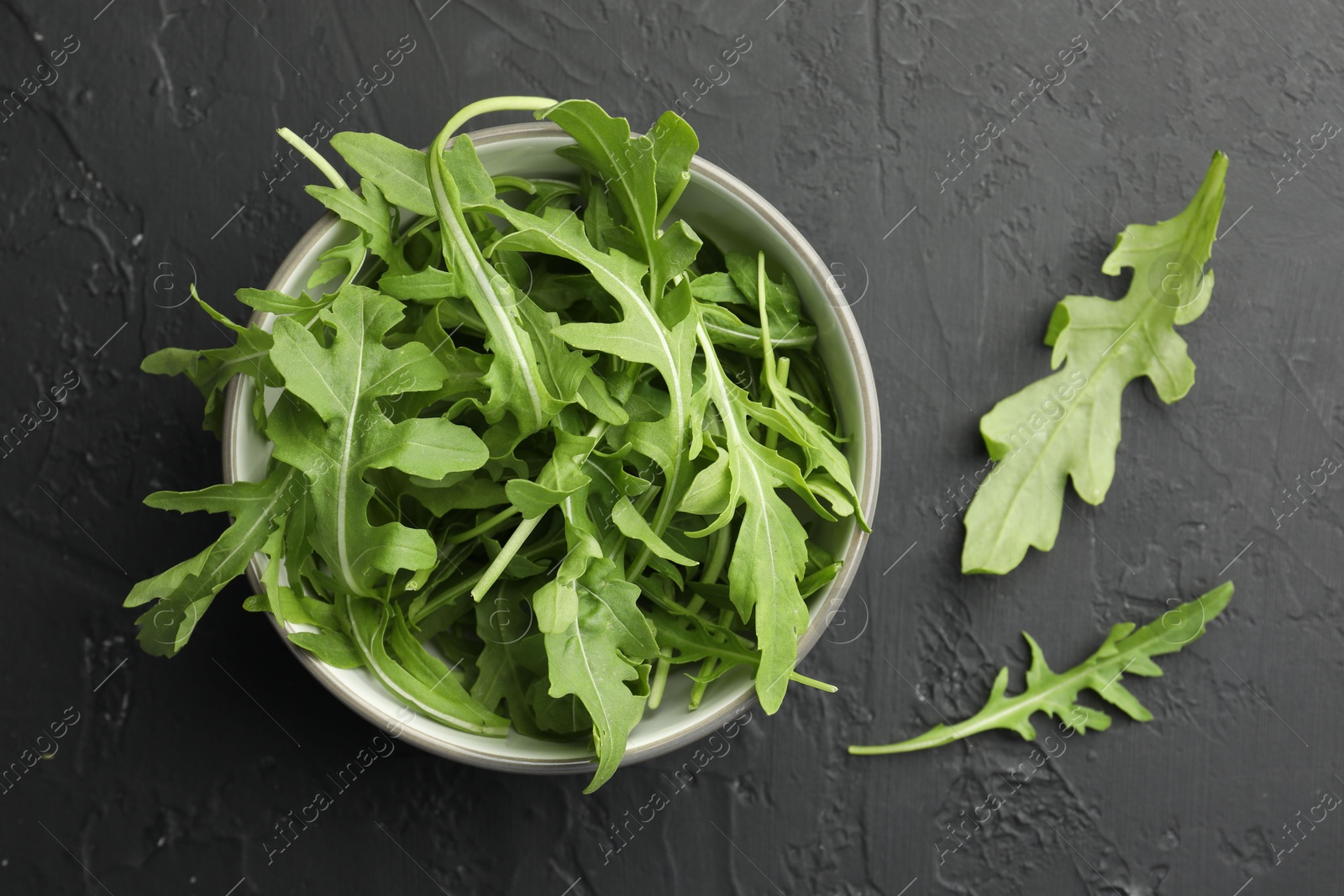 Photo of Fresh green arugula leaves in bowl on grey textured table, top view