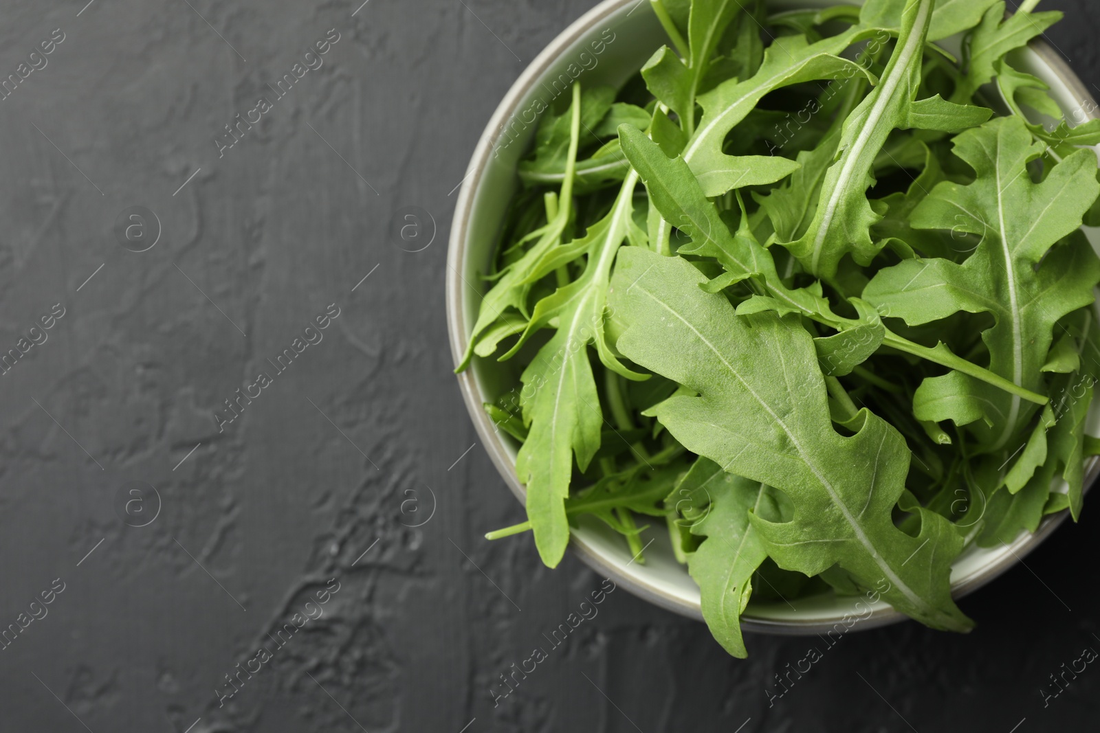 Photo of Fresh green arugula leaves in bowl on grey textured table, top view. Space for text