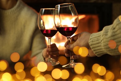 Couple clinking glasses of wine near fireplace at home, closeup. Bokeh effect