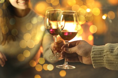 Couple clinking glasses of wine near fireplace at home, closeup. Bokeh effect