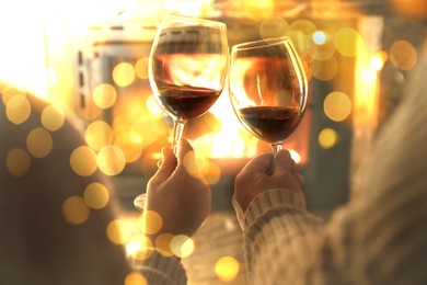 Couple clinking glasses of wine near fireplace at home, closeup. Bokeh effect