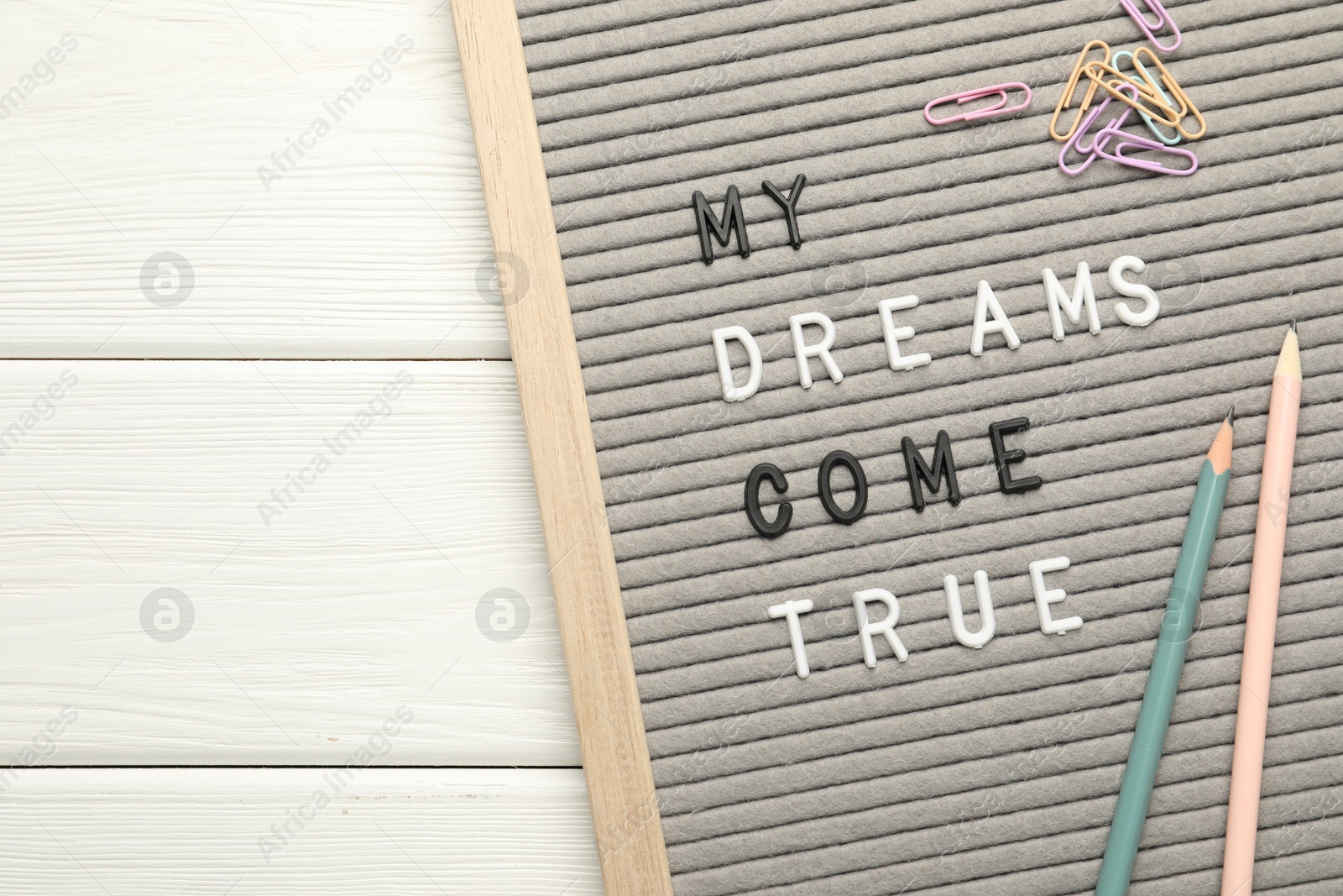 Photo of Letter board with phrase My Dreams Come True, pencils and paper clips on white wooden table, top view