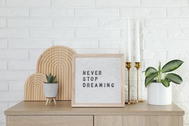 Letter board with phrase Never Stop Dreaming, plants and candles on wooden side table near white brick wall