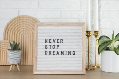 Photo of Letter board with phrase Never Stop Dreaming, plants and candles on wooden table near white brick wall