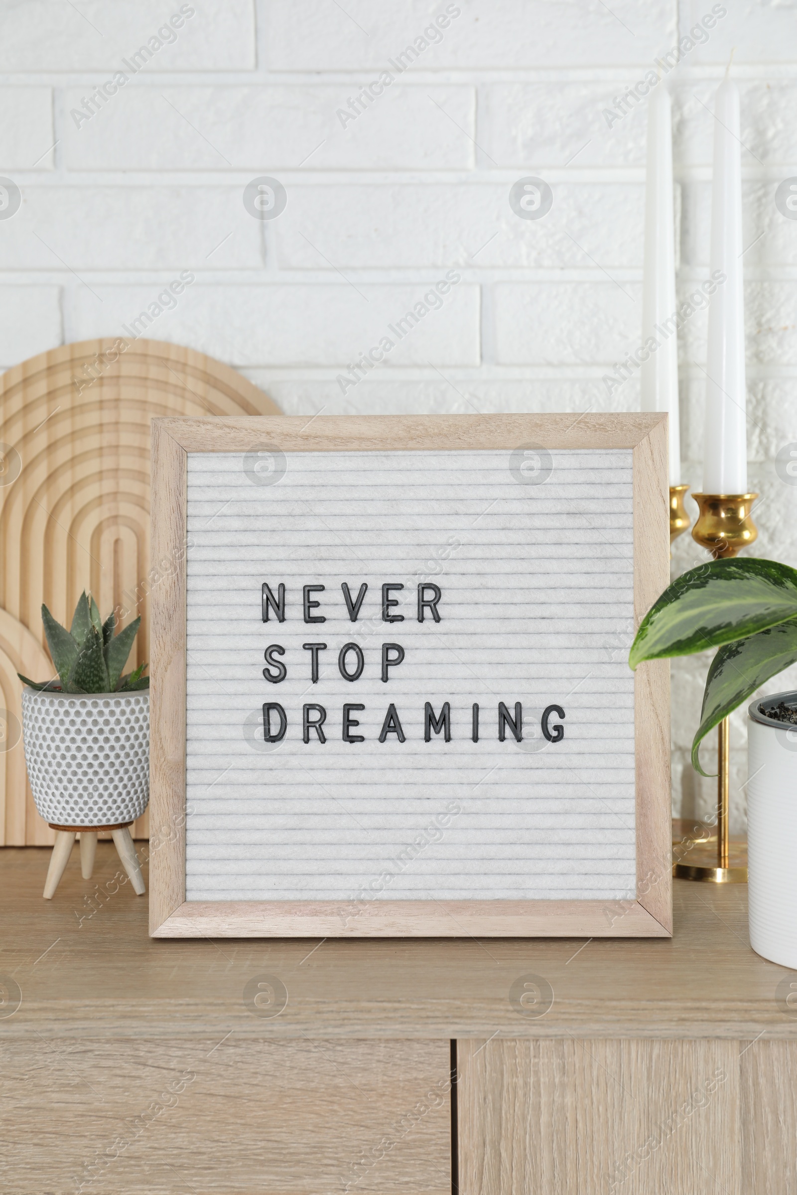 Photo of Letter board with phrase Never Stop Dreaming, plants and candles on wooden side table near white brick wall