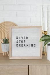 Letter board with phrase Never Stop Dreaming, plants and candles on wooden side table near white brick wall