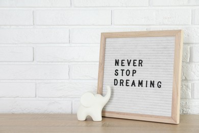 Letter board with phrase Never Stop Dreaming and elephant figure on wooden table near white brick wall, space for text