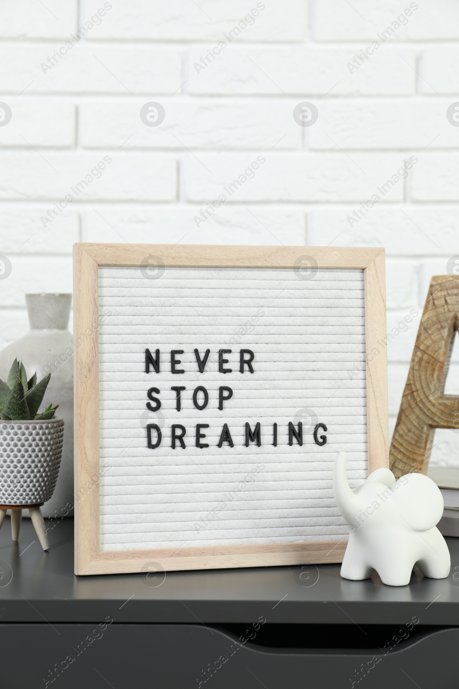 Photo of Letter board with phrase Never Stop Dreaming, plant and decor elements on grey side table near white brick wall