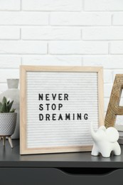 Letter board with phrase Never Stop Dreaming, plant and decor elements on grey side table near white brick wall