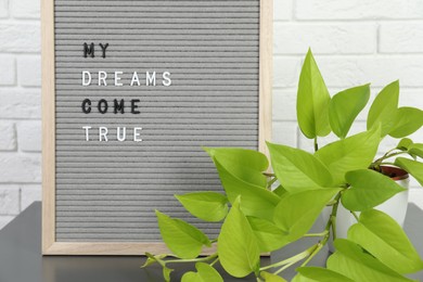 Photo of Letter board with phrase My Dreams Come True and plant on side table near white brick wall