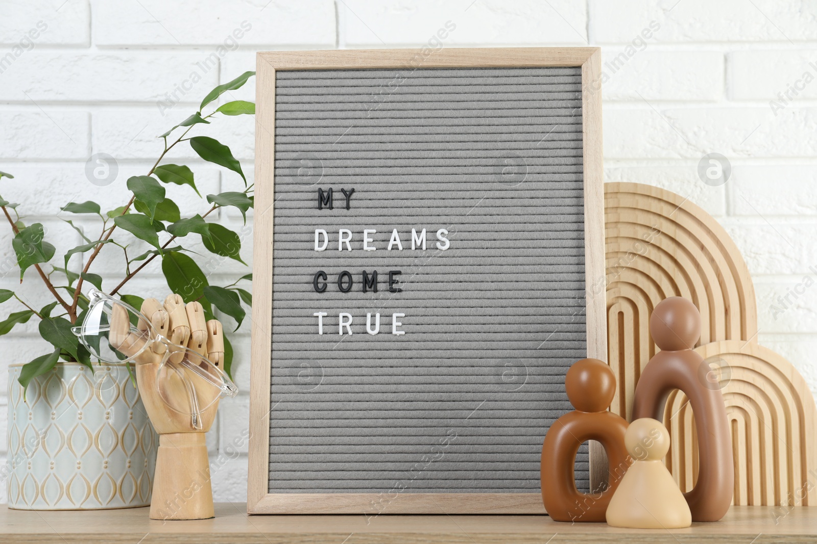 Photo of Letter board with phrase My Dreams Come True, plant and decor elements on wooden table near white brick wall