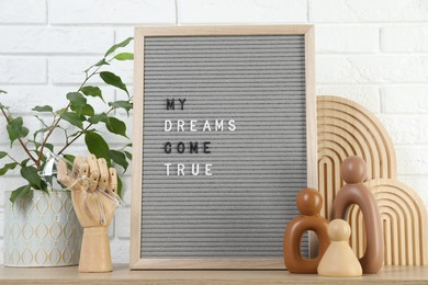 Letter board with phrase My Dreams Come True, plant and decor elements on wooden table near white brick wall