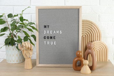 Letter board with phrase My Dreams Come True, plant and decor elements on wooden table near white brick wall