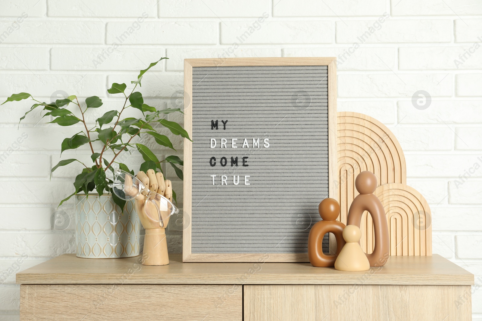 Photo of Letter board with phrase My Dreams Come True, plant and decor elements on wooden side table near white brick wall