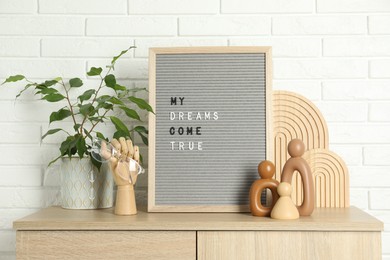 Letter board with phrase My Dreams Come True, plant and decor elements on wooden side table near white brick wall