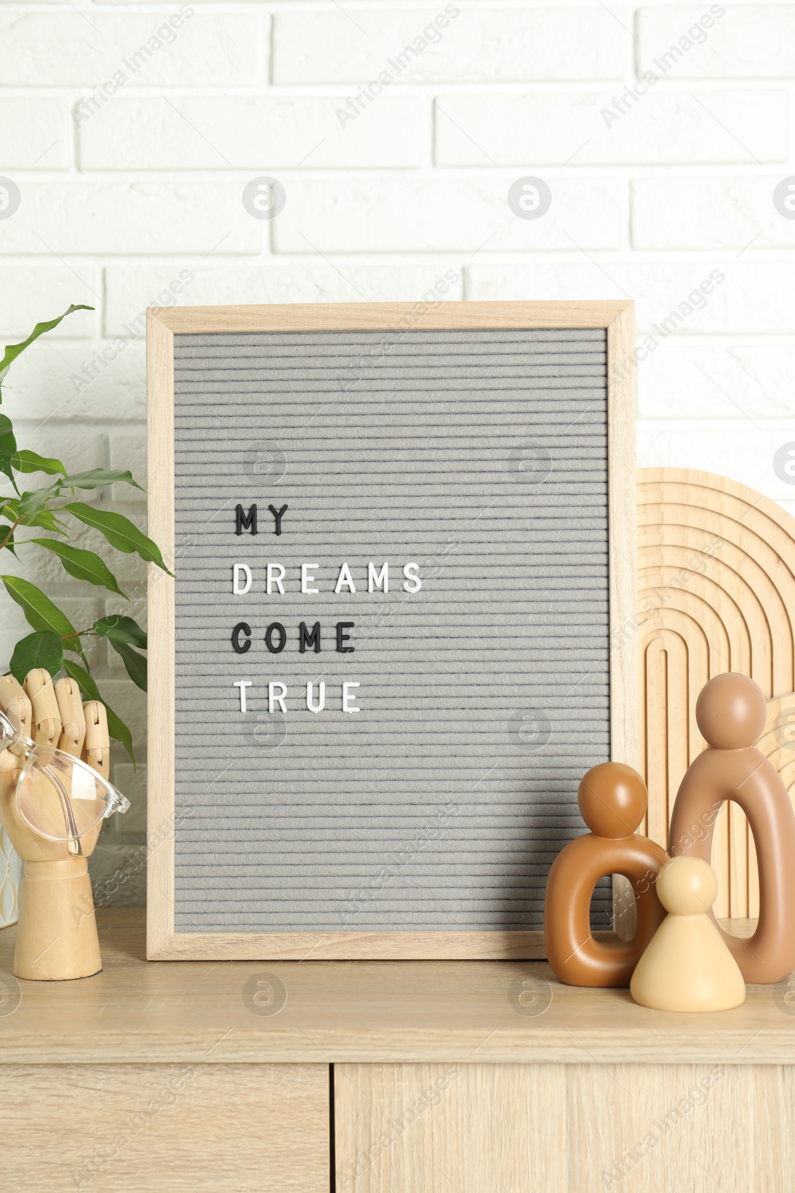 Photo of Letter board with phrase My Dreams Come True, plant and decor elements on wooden side table near white brick wall