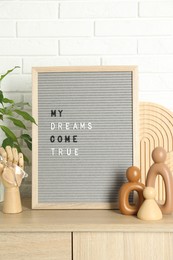 Letter board with phrase My Dreams Come True, plant and decor elements on wooden side table near white brick wall