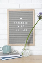 Letter board with phrase Remember Who You Are, books, cup and flower in vase on wooden table indoors