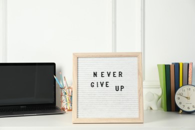 Letter board with phrase Never Give Up, laptop, alarm clock and stationery on white desk