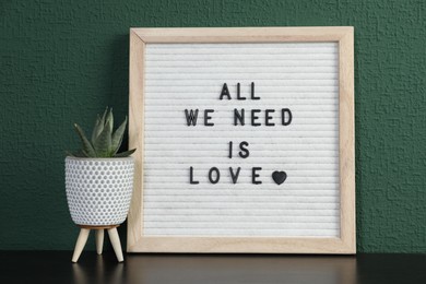 Letter board with phrase All We Need Is Love and plant on table near green wall