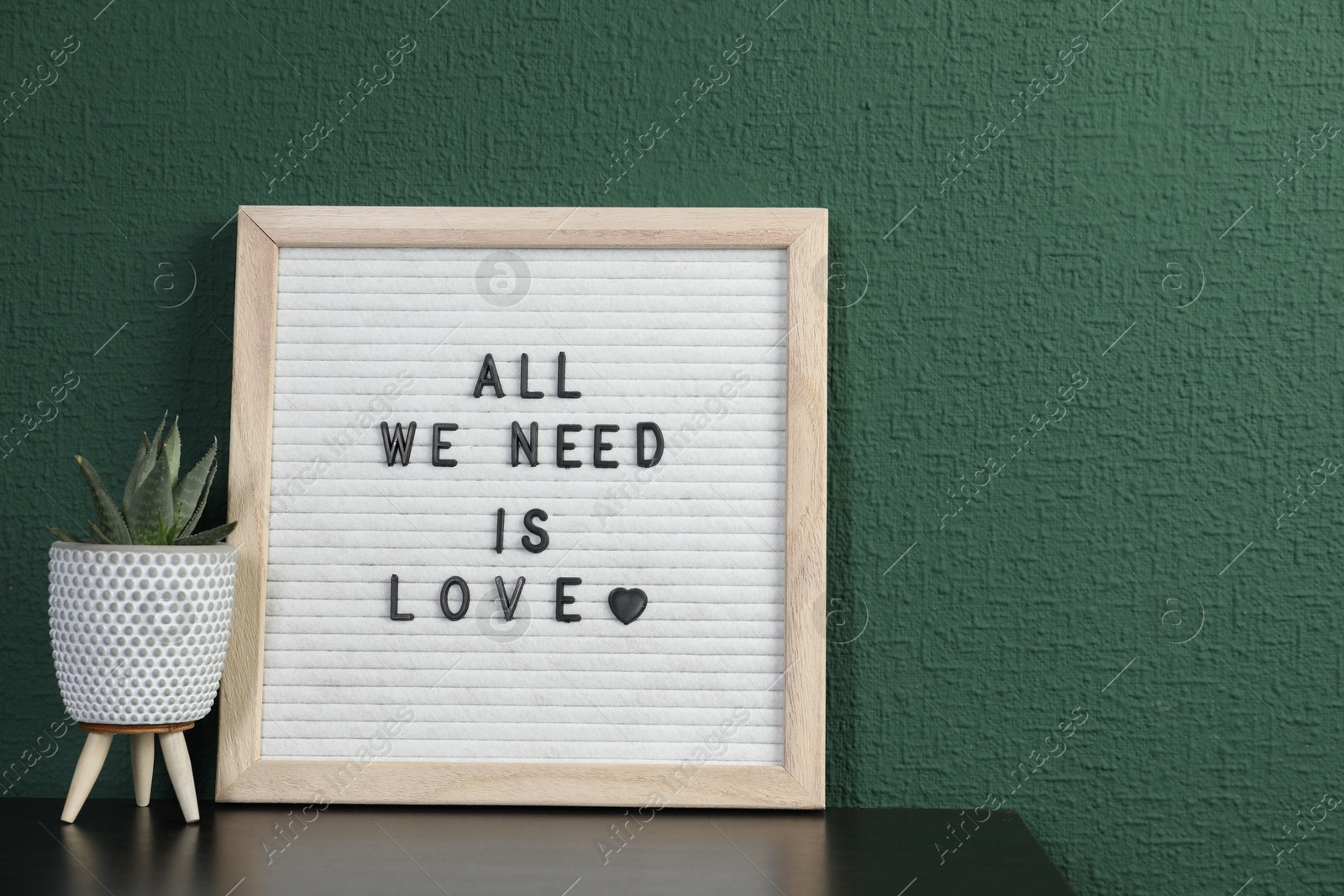 Photo of Letter board with phrase All We Need Is Love and plant on table near green wall. Space for text