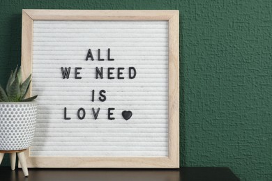 Photo of Letter board with phrase All We Need Is Love and plant on table near green wall. Space for text